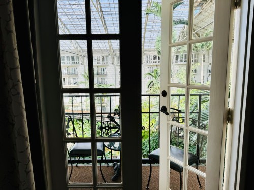 A view from inside a room showing a balcony with two chairs and a table, surrounded by greenery and glass architecture.