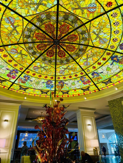 Colorful stained glass ceiling with floral and geometric patterns above a decorative glass sculpture in a hotel lobby.