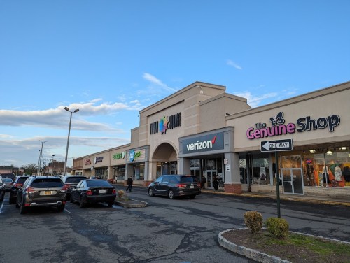 A shopping plaza with various stores, including Verizon and The Genuine Shop, under a clear blue sky.