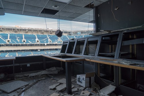 An abandoned stadium press box with broken windows, debris, and empty seats visible in the background.