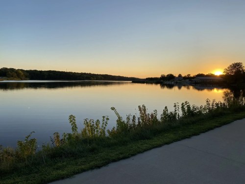 Sunset over a calm lake, with trees and grass along the shore, reflecting warm colors in the water.
