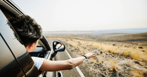 A person's arm hangs out of a car window on a road trip, with a vast, arid landscape in the background.