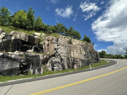A winding road curves around a rocky cliff with greenery on top under a blue sky with scattered clouds.