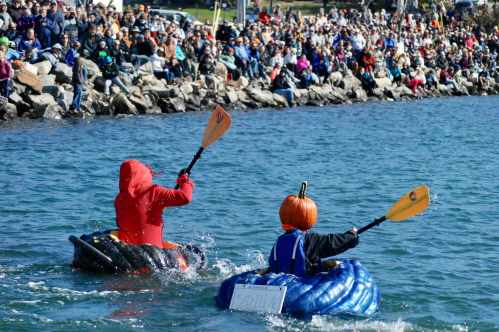 Two people in pumpkin-themed boats paddle on a lake, with a large crowd watching from the shore.