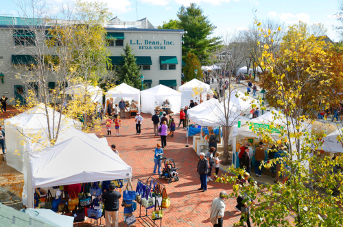 A bustling outdoor market with white tents, shoppers, and colorful fall foliage near an L.L. Bean retail store.