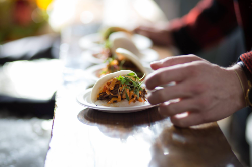 A hand reaches for a plate of tacos filled with colorful toppings on a wooden table.