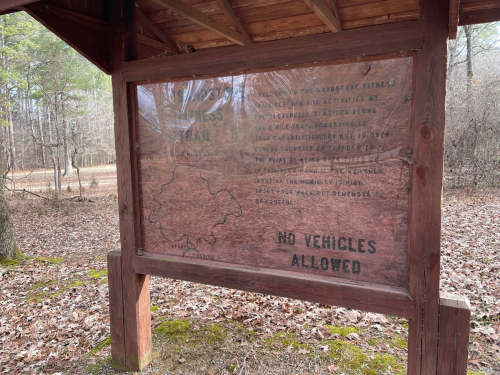 A wooden sign under a shelter detailing trail rules and a map, surrounded by trees and fallen leaves. No vehicles allowed.