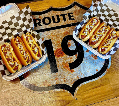 Two trays of hot dogs with mustard and onions on a rustic Route 19 sign background.