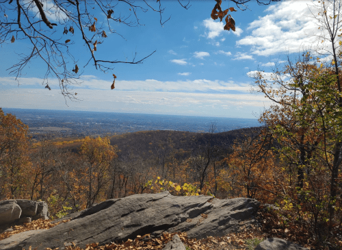 A scenic view from a mountain, showcasing autumn foliage and a vast landscape under a partly cloudy sky.