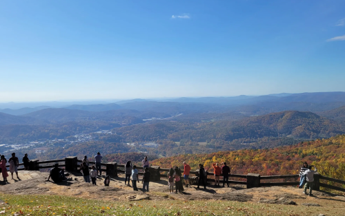 A scenic view from a mountain overlook, with people enjoying the landscape of colorful autumn trees and distant hills.