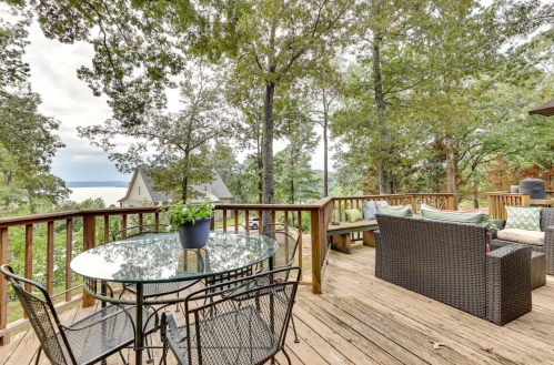 A wooden deck with a glass table and seating, surrounded by trees and a view of a lake in the background.