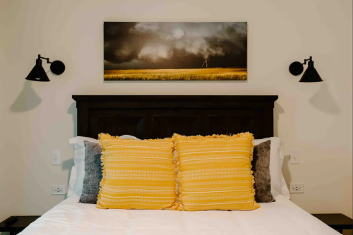 A cozy bedroom featuring a dark wooden headboard, yellow decorative pillows, and a landscape painting above the bed.