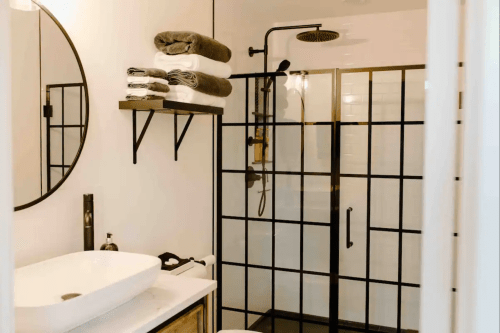 Modern bathroom featuring a glass shower, round mirror, and neatly stacked towels on a shelf.