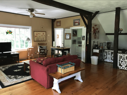 Cozy living room with a red sofa, wooden floors, a TV, and a dining area featuring rustic decor and natural light.