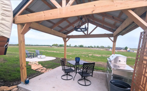 Outdoor patio with a grill, table, and chairs, overlooking a grassy field and distant trees.
