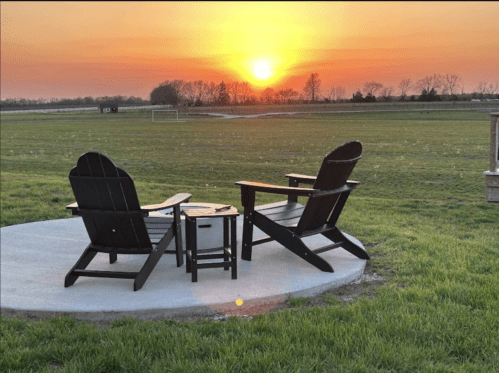 Two black Adirondack chairs and a small table on a patio, overlooking a sunset over a grassy field.