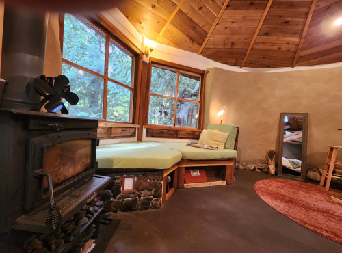 Cozy interior of a round cabin with a wood stove, large windows, and a comfortable bed nook. Natural decor and warm lighting.