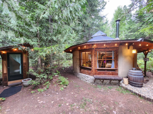 A cozy round cabin with a wooden porch, surrounded by trees and string lights, next to a small door and a barrel.