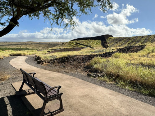 A winding path leads to a bench, surrounded by grassy fields and distant hills under a partly cloudy sky.