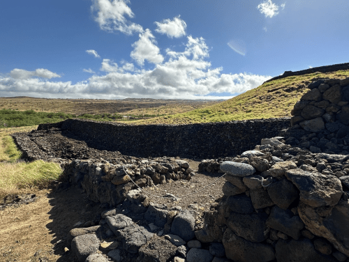 Stone walls of an ancient structure against a backdrop of rolling hills and a blue sky with scattered clouds.