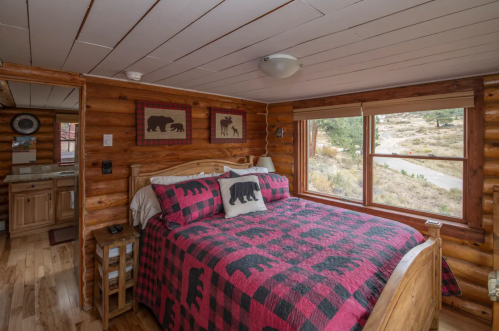 Cozy log cabin bedroom with a bear-themed quilt, wooden decor, and large windows overlooking a natural landscape.
