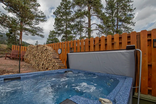 A hot tub surrounded by trees and a wooden fence, with a stack of firewood nearby under a cloudy sky.