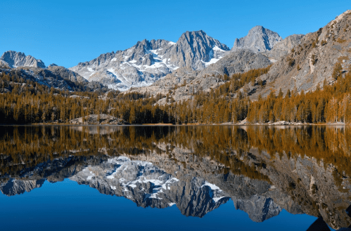 A serene mountain landscape with snow-capped peaks reflecting in a calm lake, surrounded by autumn-colored trees.