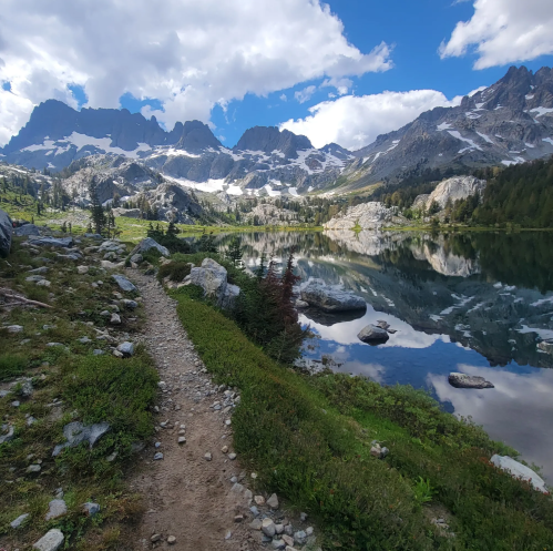 A scenic mountain landscape with a clear lake, rocky path, and lush greenery under a partly cloudy sky.