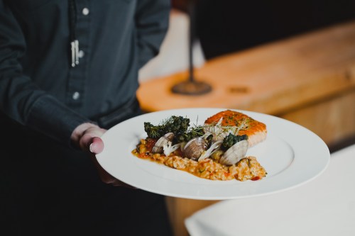 A server presents a plate with salmon, clams, and broccolini on a bed of risotto, garnished with herbs.