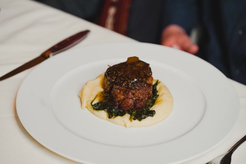 A plated dish featuring a beef filet on a bed of greens, topped with sauce, served with creamy mashed potatoes.