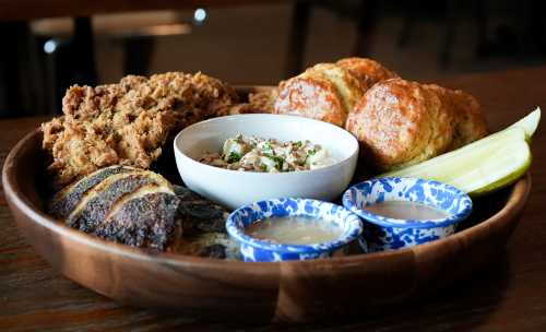 A wooden platter with fried chicken, biscuits, coleslaw, pickles, and two sauces in blue patterned bowls.