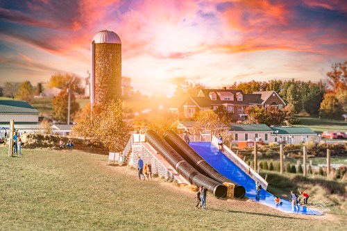 A vibrant sunset over a farm with a large slide, people enjoying the outdoors, and a barn in the background.