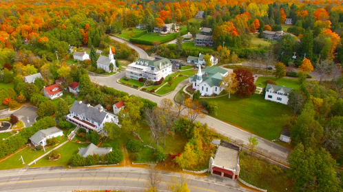 Aerial view of a picturesque village with colorful autumn foliage, featuring homes, churches, and winding roads.