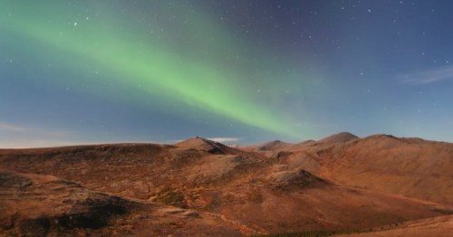 Aurora Borealis (Northern Lights) glowing green over rolling, brown alpine tundra hills under a clear, starry night sky.