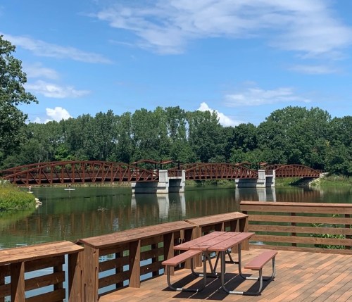 A wooden deck with a picnic table overlooks a bridge spanning a calm river, surrounded by lush greenery and blue skies.