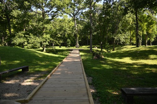 A wooden pathway leads through a lush green park, surrounded by trees and grassy mounds.