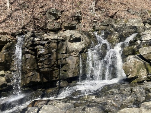 A serene waterfall cascading over rocky terrain, surrounded by dry leaves and trees in a natural setting.