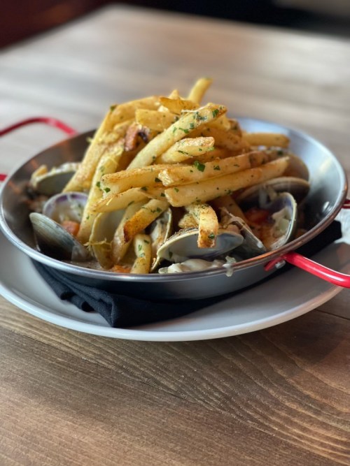 A metal dish filled with clams and topped with golden French fries, garnished with herbs on a wooden table.