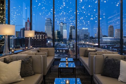 A stylish lounge with modern sofas and tables, featuring a panoramic view of a city skyline under a starry night sky.