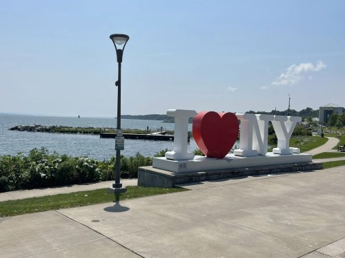 A large "I ❤️ NY" sign by the waterfront, with a clear blue sky and a calm lake in the background.