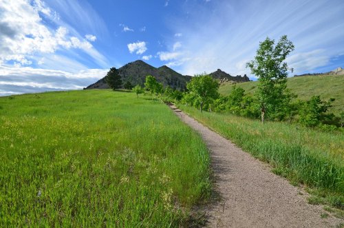 A winding dirt path through lush green grass, leading towards a mountain under a blue sky with wispy clouds.