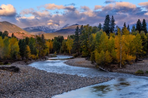 A winding river flows through a forest of autumn trees, with snow-capped mountains and clouds in the background.