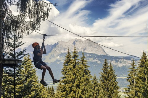 A person in a helmet zip-lining through trees with mountains and a blue sky in the background.