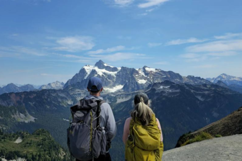 Two hikers with backpacks stand on a trail, gazing at a snow-capped mountain range under a clear blue sky.