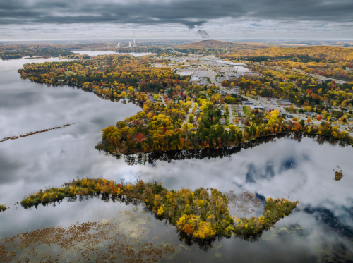 Aerial view of a colorful autumn landscape with a winding river, trees, and a distant industrial area under cloudy skies.