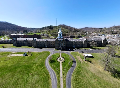 Aerial view of a historic building with a clock tower, surrounded by green lawns and hills in the background.