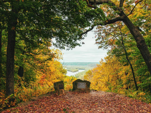 Scenic view from a forested overlook, featuring autumn foliage and a river in the distance. Two stone markers in foreground.