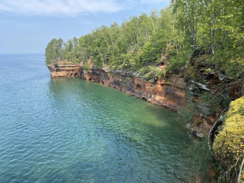 A serene view of a rocky shoreline with lush green trees and calm blue water under a clear sky.