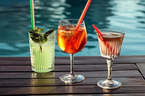 Three colorful cocktails with straws sit on a wooden table by a shimmering pool.