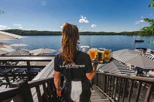 A server carries drinks down wooden steps towards a lakeside patio with striped umbrellas and a scenic view.